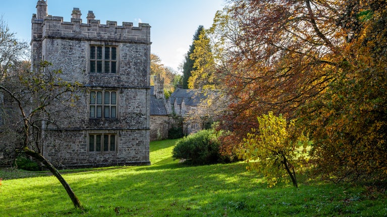 Exterior view of grey stone building, seen across a green meadow. Trees with orange and brown leaves on right side of image.